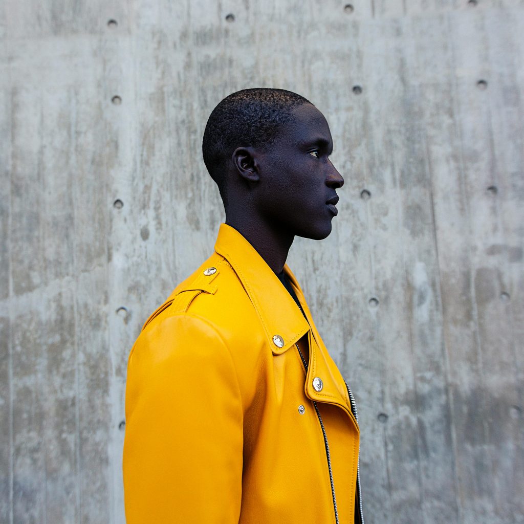 A fashion-forward portrait of a man in a vibrant yellow jacket against a concrete backdrop.