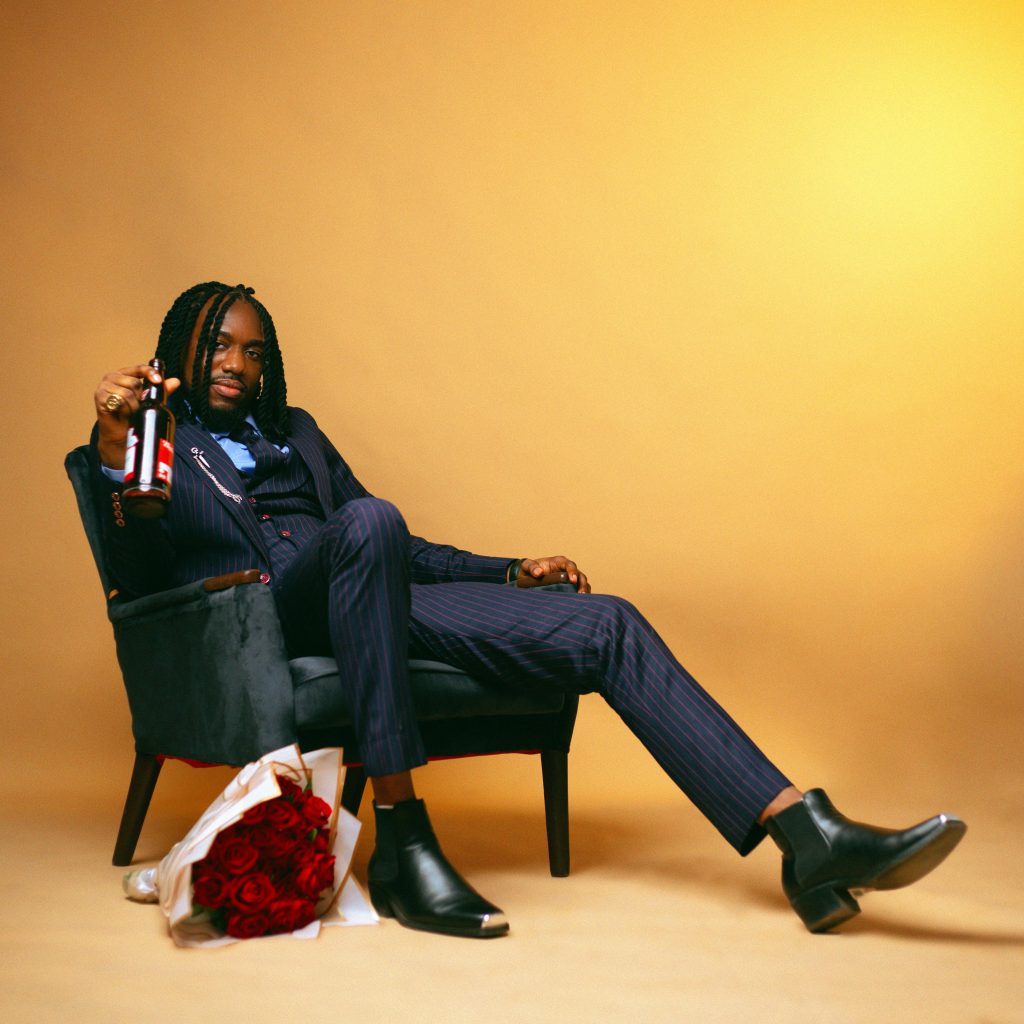 Man in a suit lounging with a beer bottle and roses, set against a warm background.