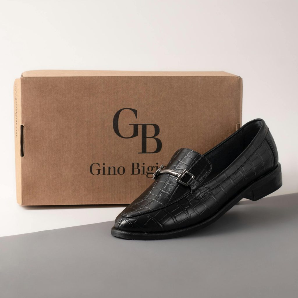 Black leather shoe with buckle detail next to branded Gino Bigioni box.