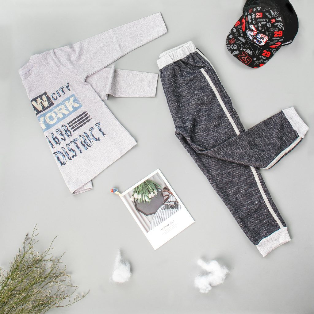 Flat lay of children's casual outfit with shirt, pants, and cap on gray background.